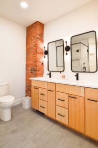 View of the double vanity in an updated bathroom with an exposed brick wall and modern black and white tiled shower.