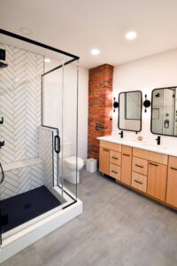 View of the double vanity in an updated bathroom with an exposed brick wall and modern black and white tiled shower.