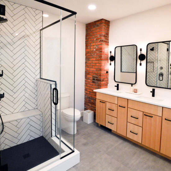 View of the double vanity in an updated bathroom with an exposed brick wall and modern black and white tiled shower.