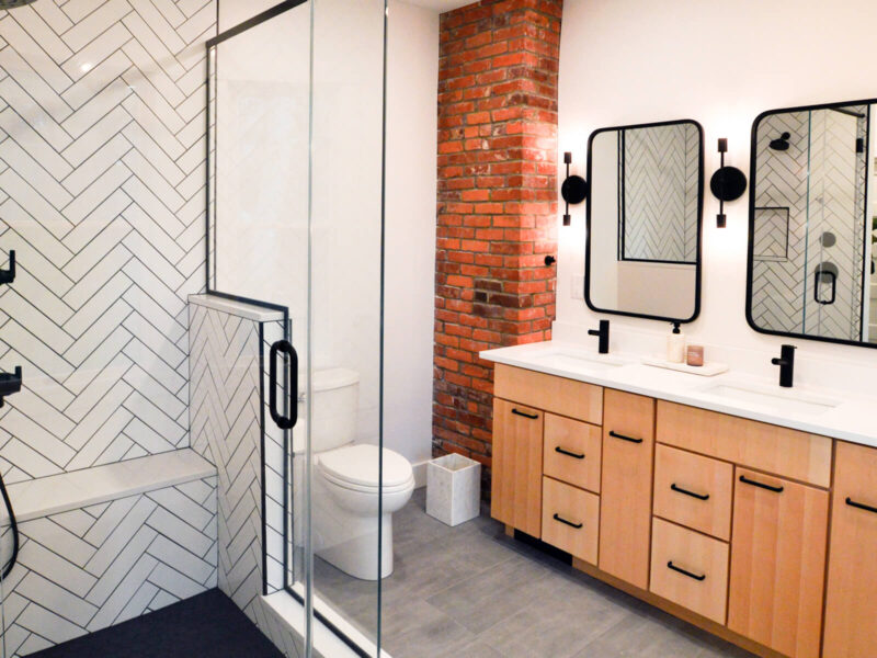 View of the double vanity in an updated bathroom with an exposed brick wall and modern black and white tiled shower.