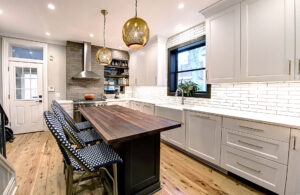 View of the two toned island in a newly remodeled kitchen featuring a large natural wood island, exposed brick, black windows, and pendant lights.