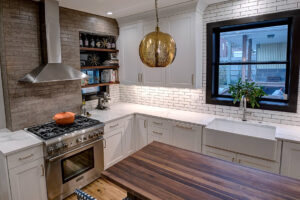 View of a newly remodeled kitchen featuring two-toned cabinetry, a large natural wood island, exposed brick, black windows, and pendant lights.