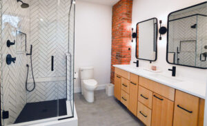 View of a newly expanded bathroom featuring warm earth tones, an exposed brick wall, modern black and white tiled shower, and tons of natural light.