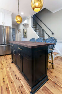 View of the black island with a natural wood countertop in a newly remodeled kitchen featuring two-toned cabinetry, exposed brick, black windows, and pendant lights.