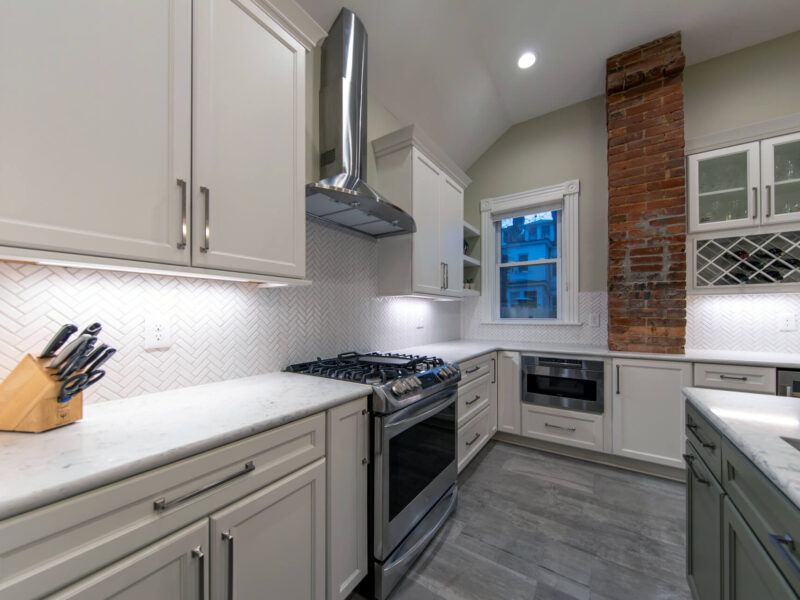 View of a remodeled classic white kitchen with marble countertops, herringbone tile backsplash, and built-in wine storage.