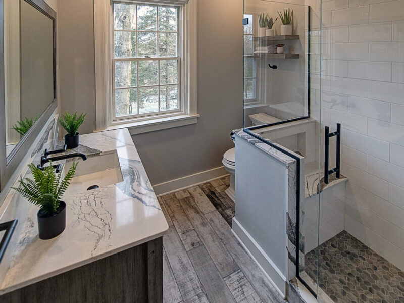 Broad view of an updated bathroom with a vanity with a marble countertop and weathered wood flooring.