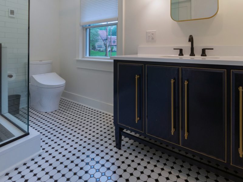 View of a remodeled bathroom with a classic, black and white checkered tile floor, walk-in shower with glass walls, and a large vanity.