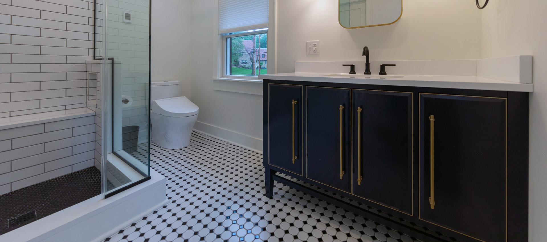 View of a remodeled bathroom with a classic, black and white checkered tile floor, walk-in shower with glass walls, and a large vanity.