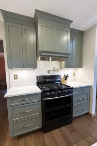 Close up view of the black stove and green cabinets with matching wood vent hood.