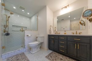 Broad view of a master bathroom with white subway tiled shower, marble accents, a double vanity, and gold hardware.