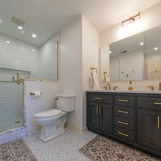 Broad view of a master bathroom with white subway tiled shower, marble accents, a double vanity, and gold hardware.
