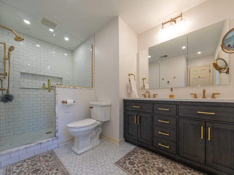 Broad view of a master bathroom with white subway tiled shower, marble accents, a double vanity, and gold hardware.