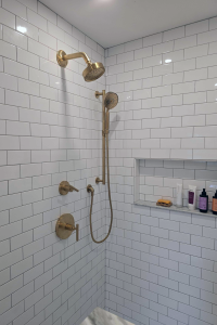 Close up view of the gold shower hardware in a remodeled bathroom. Shower features white subway tile and marble accents.