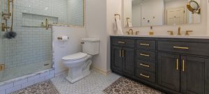 Broad view of a master bathroom with white subway tiled shower, marble accents, a double vanity, and gold hardware.