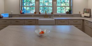 View of a newly remodeled kitchen looking over the huge island with marble counter and farmhouse sink with three windows in the background.