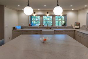 View of a newly remodeled kitchen looking over the huge island with marble counter and farmhouse sink with three windows in the background.