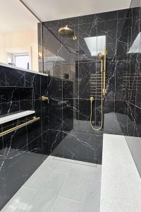 Close up view of a walk-in shower with black tile, gold fixtures, and a hidden drain.